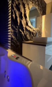 3D lávakő sziklafal modern fürdőszobában, kör alakú tükörrel és rejtett világítással. Kézzel formált kőhatású falburkolat, amely exkluzív és prémium megjelenést biztosít. A luxus fürdőszobát hidromasszázs kád egészíti ki, kék LED fényekkel. Országos sziklafal kivitelezés – egyedi, természetközeli dizájn otthonokba, hotelekbe és wellness terekbe.