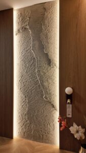 Exkluzív 3D Fillit sziklafal textúra: kézzel formált bézs kőhatású dekoráció modern étkezőkbe vagy nappali fókuszfalnak, elegáns súrolófényekkel és természetes fa panelekkel keretezve.